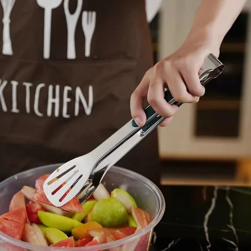 Person wearing a brown apron with kitchen utensils holding tongs over a container of fruit salad.